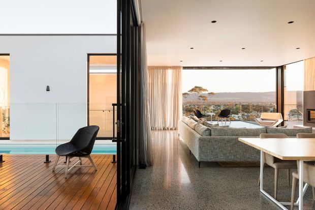 A contemporary open-plan living and dining area opening onto a timber deck and pool terrace through expansive black-framed glazed doors. The interior features a large textured sectional sofa, dining table, polished concrete or terrazzo-like flooring, sheer curtains, recessed ceiling lights, and broad views across a suburban landscape. A sculptural lounge chair sits on the deck beside the pool.