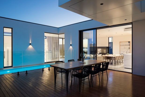 A contemporary outdoor dining terrace with dark timber decking, a long dining table and chairs, and a narrow illuminated water feature running along a blue rendered wall. Large glazed openings connect the terrace to a bright open-plan interior with a dining area and kitchen. The space features clean geometric lines, recessed lighting, and a projecting flat roof canopy.