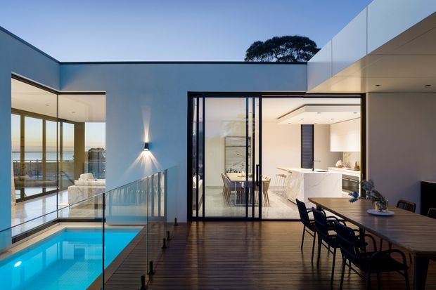 A contemporary flat-roofed home with a timber deck overlooking a vivid blue pool. The image shows an outdoor dining area beside full-height glazing, with a bright white kitchen visible through open glass doors. Glass balustrading, clean geometric lines, and a waterfront or coastal outlook create an open, refined atmosphere.