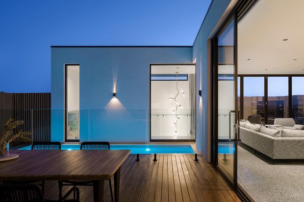 Contemporary residential terrace at dusk, with a timber deck and outdoor dining table beside a narrow illuminated swimming pool. Floor-to-ceiling glazing opens into a bright living room with a large sectional sofa, while the exterior walls are rendered in a cool blue-grey tone and framed by dark metal window and door systems.
