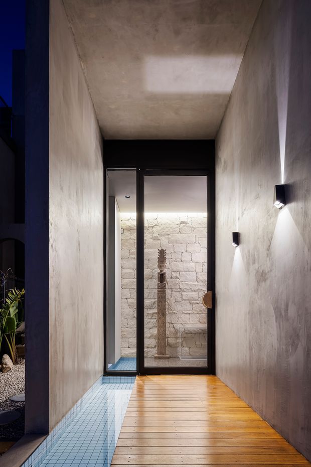 A narrow, atmospheric passage with textured plaster or concrete walls and ceiling, black-framed glazed doors, a timber walkway, and a narrow blue-tiled water channel along the floor. The glazing opens toward a stone-walled courtyard or display area with a sculptural object. Minimal wall-mounted lights create a dramatic, moody effect.