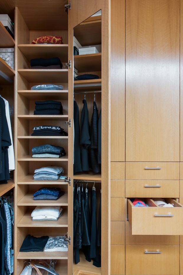 A close-up view of a custom timber walk-in wardrobe with open shelving, folded clothing, hanging garments, mirrored cabinet doors, and integrated drawers. The warm wood joinery fills the frame and provides highly organized storage.