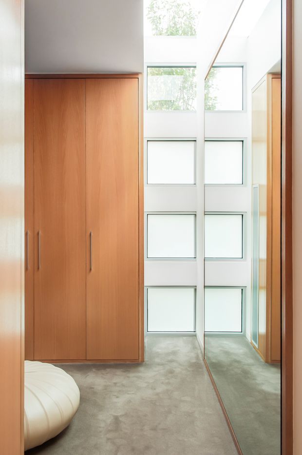 A bright, narrow wardrobe area with full-height warm timber built-in cabinetry, long vertical handles, plush light grey carpeting, and a large full-height mirror reflecting the cabinetry and windows. White-framed stacked windows bring in abundant natural light, creating a clean and airy interior.