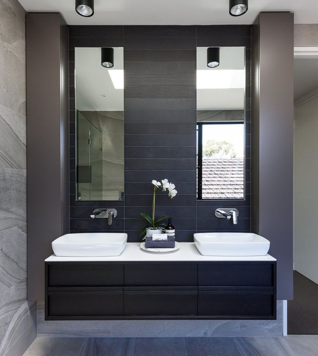 Contemporary double-vanity bathroom with two white vessel basins on a dark built-in floating vanity. The room features dark textured wall tiles, large mirrored cabinets, chrome wall-mounted tapware, recessed and surface-mounted lighting, and large-format grey stone-look wall and floor finishes. A narrow window brings in daylight and reflects the roof outside.