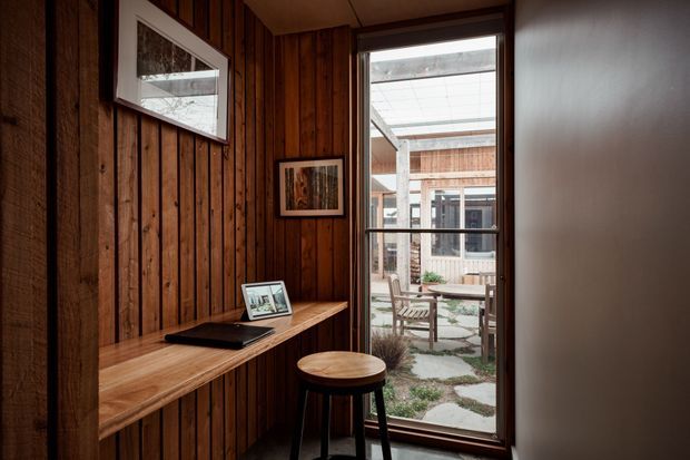 A compact timber-lined interior nook with a built-in wooden desk or console, laptop, stool, framed artwork, and a full-height glazed door or window opening onto a small courtyard. The space has warm, dark natural timber panelling and a simple contemporary cabin-like character.