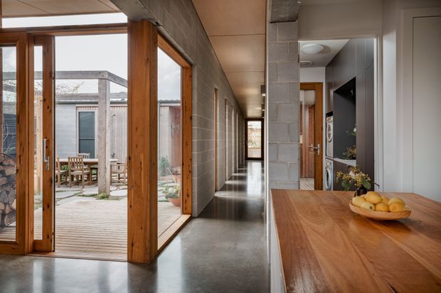 A long, narrow contemporary interior corridor with polished concrete flooring, exposed grey concrete-block walls, warm timber-framed glazing and doors, and a timber kitchen island or counter in the foreground. Full-height glazing opens onto a small timber deck and courtyard, while a wall-mounted television and built-in cabinetry are visible on the right. The space combines robust industrial materials with warm timber detailing and abundant natural light.