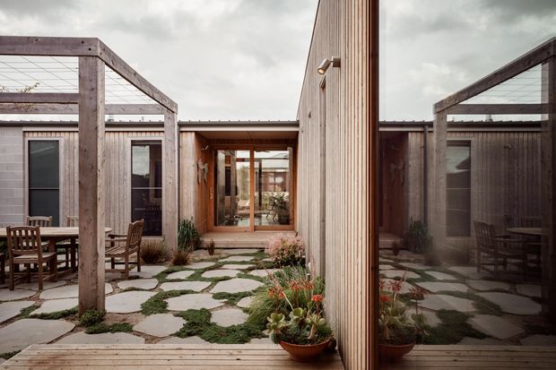 A contemporary courtyard viewed from an outdoor timber deck, featuring irregular stone paving set among groundcover planting, timber pergola structures, vertical timber cladding, large reflective glazing, and warm timber-framed openings. Outdoor dining furniture sits beneath the pergola, while a planted pot and reflected courtyard add depth. The overcast sky creates a muted, calm atmosphere.