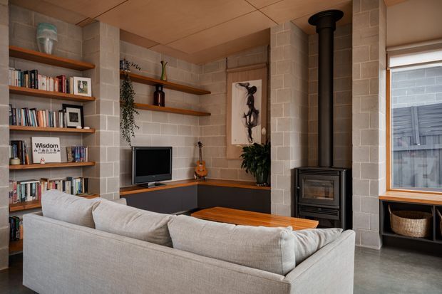A warm, compact living room with built-in open bookshelves lining the left wall, a low dark media cabinet and television, a corner wood-burning stove, and a light upholstered sectional sofa in the foreground. Exposed grey concrete-block walls, timber shelving and ceiling panels, a timber-framed window, and polished grey flooring create a rugged yet comfortable interior. A small violin, artwork, books, and indoor plants add character.