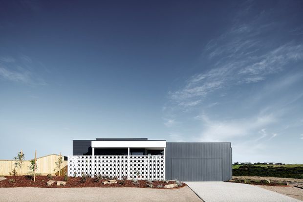 Front exterior view of a low, single-storey contemporary residence with a flat roof, dark and light rendered volumes, a distinctive white breeze-block screen across the entry or living area, and a large ribbed grey garage door. The property sits in a sparsely landscaped suburban setting with a broad paved driveway and open sky.