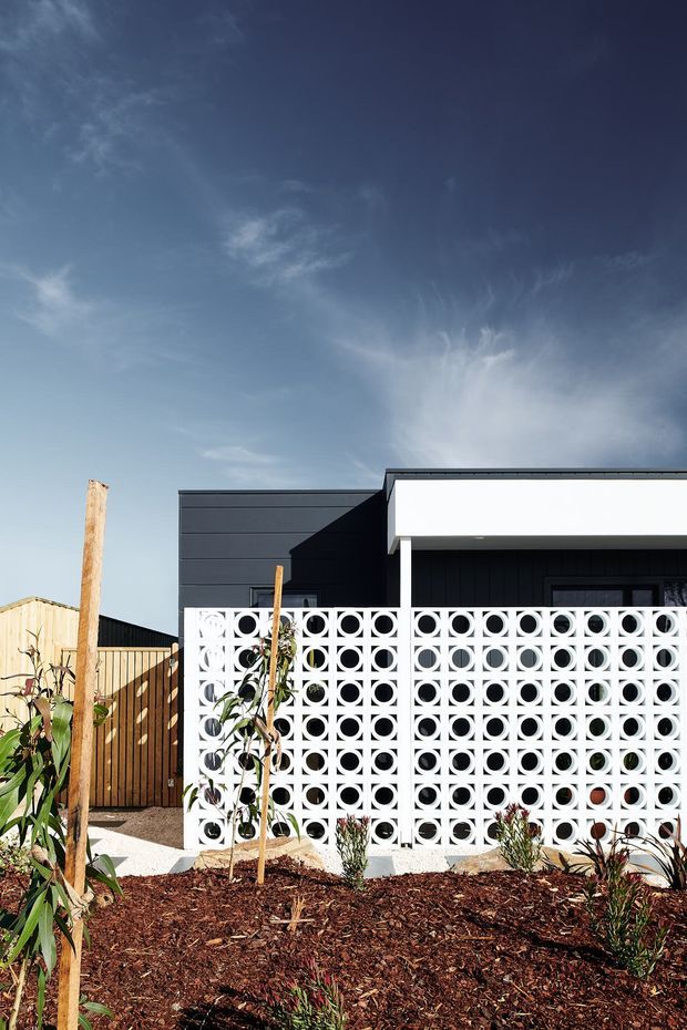 Exterior view of a contemporary single-storey building with dark horizontal cladding, a crisp white roof-edge canopy, and a prominent white breeze-block screen forming a patterned semi-enclosed frontage. A newly landscaped garden bed with mulch and young plants occupies the foreground.