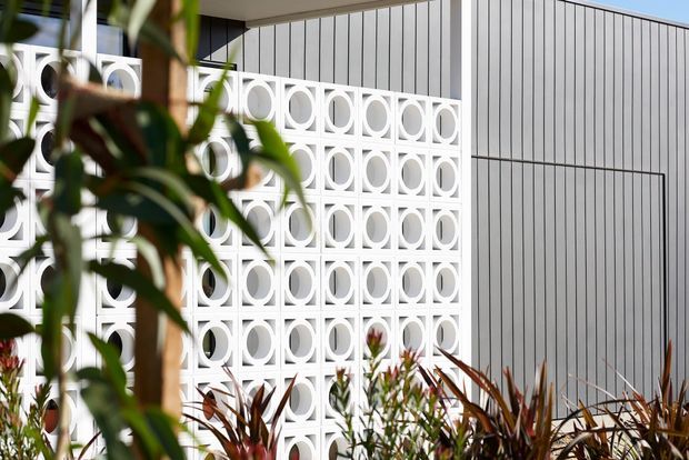 Exterior architectural view featuring a white geometric breeze-block screen with repeating circular cut-outs beside vertically ribbed light-grey cladding. The facade is partially framed by tropical and ornamental planting, with a flat, modern roofline and strong sunlight.