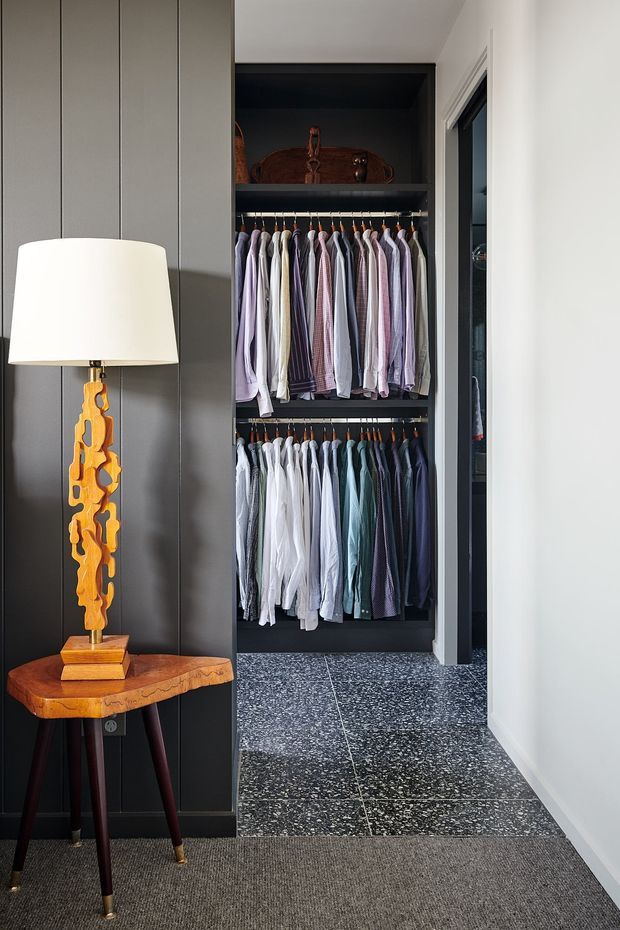 A refined walk-in wardrobe and dressing area with open dark cabinetry, two rows of neatly arranged shirts, and a high storage shelf. The space features dark vertical wall panelling, speckled dark stone or terrazzo-style floor tiles, and a grey carpeted foreground. A sculptural amber timber table lamp sits on a small live-edge timber side table, adding a warm accent to the otherwise dark, tailored interior.