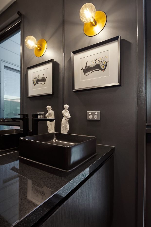 A compact, moody bathroom vanity with a glossy black vessel basin set on a dark stone benchtop and dark built-in cabinetry. Charcoal walls are decorated with framed artwork and illuminated by warm gold wall sconces. A large mirror reflects the vanity and adjacent window, while white electrical outlets and small classical figurines add detail.