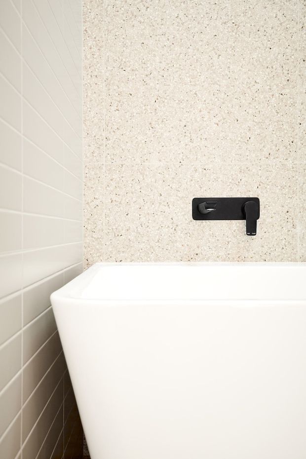 Close-up of a minimalist bathroom featuring a white freestanding bathtub, pale textured stone-look wall tiles, white rectangular wall tiles, and a matte black wall-mounted bath tap and spout.