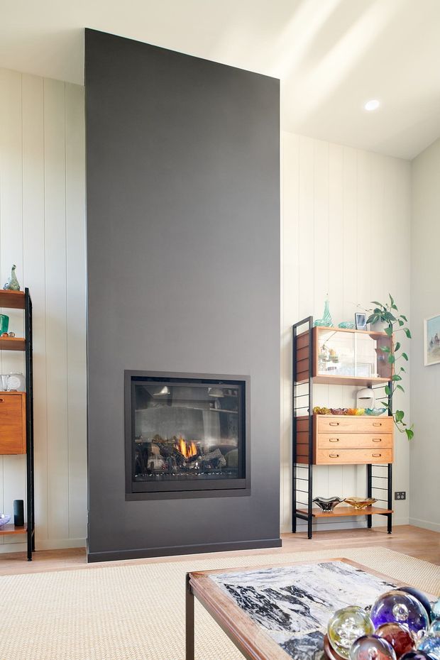 Bright contemporary living room featuring a dramatic full-height dark grey fireplace feature wall with an inset firebox, pale built-in wall cabinetry, timber open shelving and display joinery, and a marble-topped coffee table. Natural timber flooring, a light woven rug, and indoor plants add warmth to the minimalist interior.