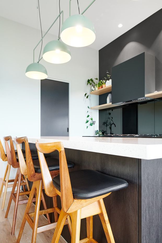 A bright contemporary kitchen featuring a long island with a white stone benchtop, dark vertically textured cabinetry, black splashback and integrated cooktop, open timber shelving with plants, and several sculptural pale green pendant lights. Warm timber bar stools with black upholstered seats line the island.
