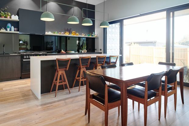 A contemporary open-plan kitchen and dining area with dark cabinetry, a long island with timber bar stools, and a dark timber dining table with matching chairs. Three pendant lights hang above the island, while large glazed sliding doors bring in abundant daylight and open to a fenced outdoor area. Light timber flooring contrasts with the black cabinetry and dark furniture.
