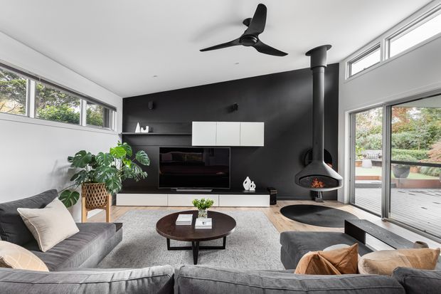 Contemporary living room with a dark feature wall, wall-mounted television and white floating cabinetry, a suspended black wood-burning fireplace, ceiling fan, large sliding glass doors, clerestory windows, grey sectional sofas, and a round timber coffee table. The glazing opens toward an outdoor deck and garden.