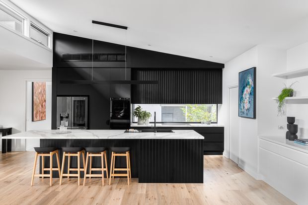 Bright contemporary kitchen with a large white stone island, four timber-and-black bar stools, and extensive black fluted cabinetry. The space has pale timber flooring, white walls and ceiling, integrated shelving, a black rangehood, pendant lighting, and clerestory-style glazing. A white built-in media or storage wall is visible at right, with artwork and plants providing restrained decorative accents.