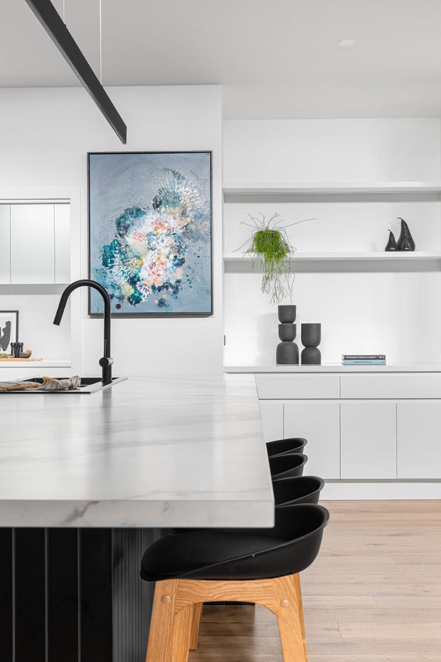 Bright contemporary kitchen with a large pale stone island, black gooseneck tap, integrated white cabinetry, open display shelving, black bar stools with timber legs, and light timber flooring. A colourful abstract artwork and simple sculptural decor add contrast, while a dark exposed ceiling beam provides a strong architectural accent.