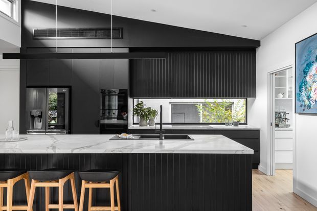 Contemporary kitchen with a large marble-look island, black vertically fluted cabinetry, integrated appliances, pendant lighting and timber bar stools. White walls and timber flooring provide contrast, while a side doorway opens toward a pantry or storage area.