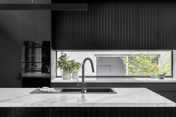 Contemporary dark-toned kitchen featuring black vertically ribbed cabinetry, integrated appliances, a large white marble-look island benchtop, double undermount sink with a curved black tap, and a wide window with black roller blinds. Potted plants and a monochrome palette add contrast and natural detail.
