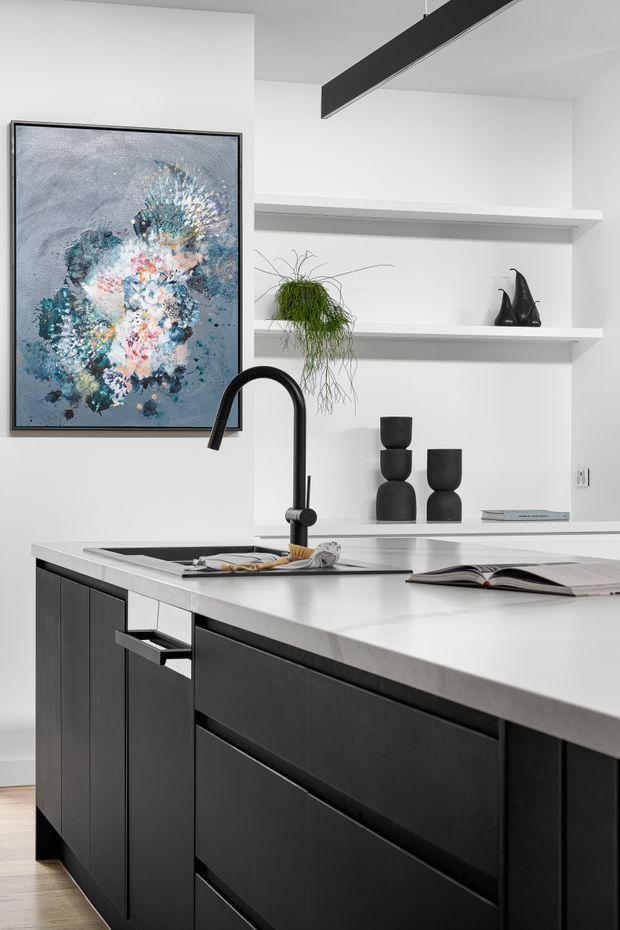 A contemporary kitchen interior featuring a large white island with an integrated black sink and curved matte-black tap, dark charcoal cabinetry with slim recessed pulls, white walls, and open display shelving. A black linear pendant light, abstract artwork, sculptural black decor, and a trailing plant add contrast and visual interest.