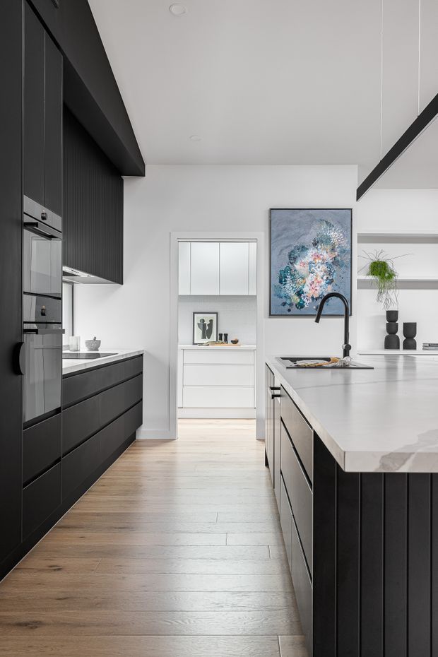 A contemporary galley-style kitchen with extensive matte black cabinetry, integrated appliances, a large white stone island with a black gooseneck tap, and light timber flooring. White walls, open shelving, recessed lighting, and a framed abstract artwork create a clean, high-contrast interior. A bright utility or pantry area is visible through the doorway at the rear.