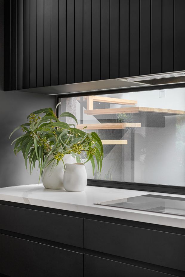 Close-up of a contemporary kitchen featuring matte black vertical-panel cabinetry and matching lower joinery, a bright white stone benchtop with an integrated dark cooktop, and a large black-framed window. A potted leafy plant sits on the benchtop, with timber stair or shelving elements visible beyond the glazing.