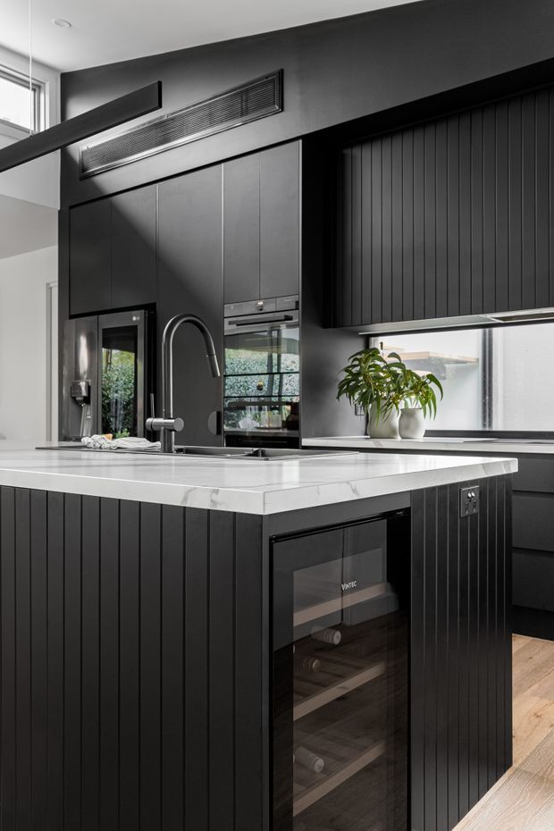 A refined contemporary kitchen viewed from the island, featuring matte black vertically fluted cabinetry, integrated black appliances, a white marble-look stone island benchtop, dark splashback and wall finishes, a sculptural black tap, and a built-in wine cooler. Natural light enters through a large horizontal window, with light timber flooring adding warmth.