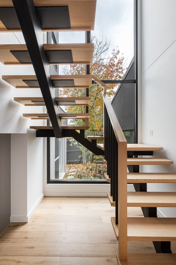 Interior view of a contemporary stairway with floating light timber treads supported by a bold black steel central spine and matching dark balusters. A timber handrail and open risers frame the staircase, while large floor-to-ceiling windows overlook a small courtyard and bring in abundant natural light. Pale timber flooring, white walls, and exposed structural elements create a clean, refined architectural composition.