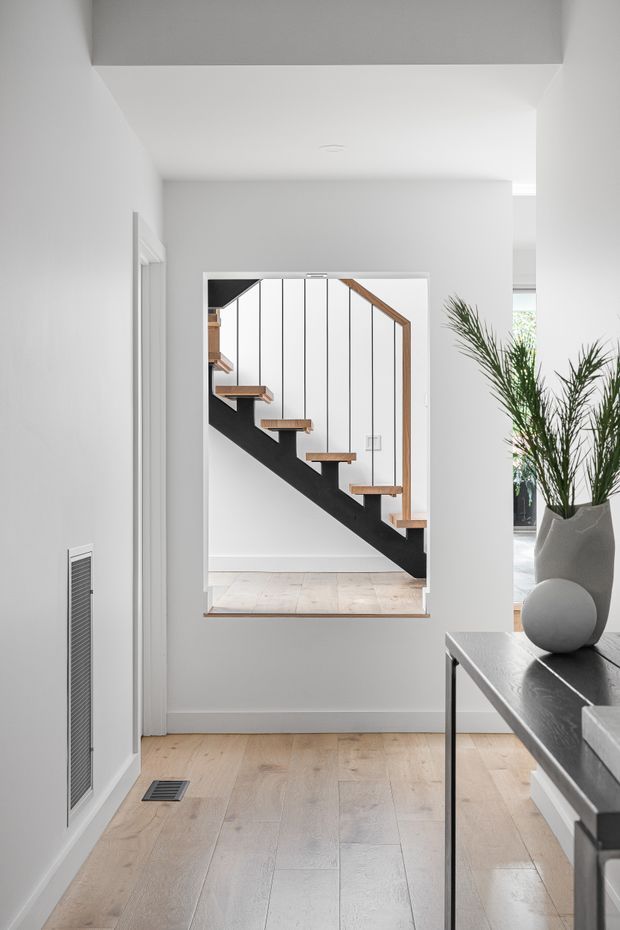 Bright minimalist interior view focused on a modern open-riser staircase seen through a large wall opening. The space has white walls and ceiling, pale timber flooring, a dark painted stair stringer, slim black vertical balusters, timber stair treads and handrail, and contemporary console furniture with a sculptural vase and tall plant. A glimpse of an adjacent bright room is visible beyond the staircase.