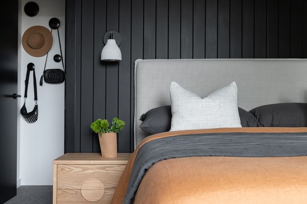 A stylish bedroom with a large upholstered light-grey bedhead against a dark vertical timber-panel feature wall. The bed is dressed in charcoal and warm tan bedding with a textured white cushion. A timber bedside cabinet, white wall sconce, potted plant, and decorative wall-mounted accessories complete the room.