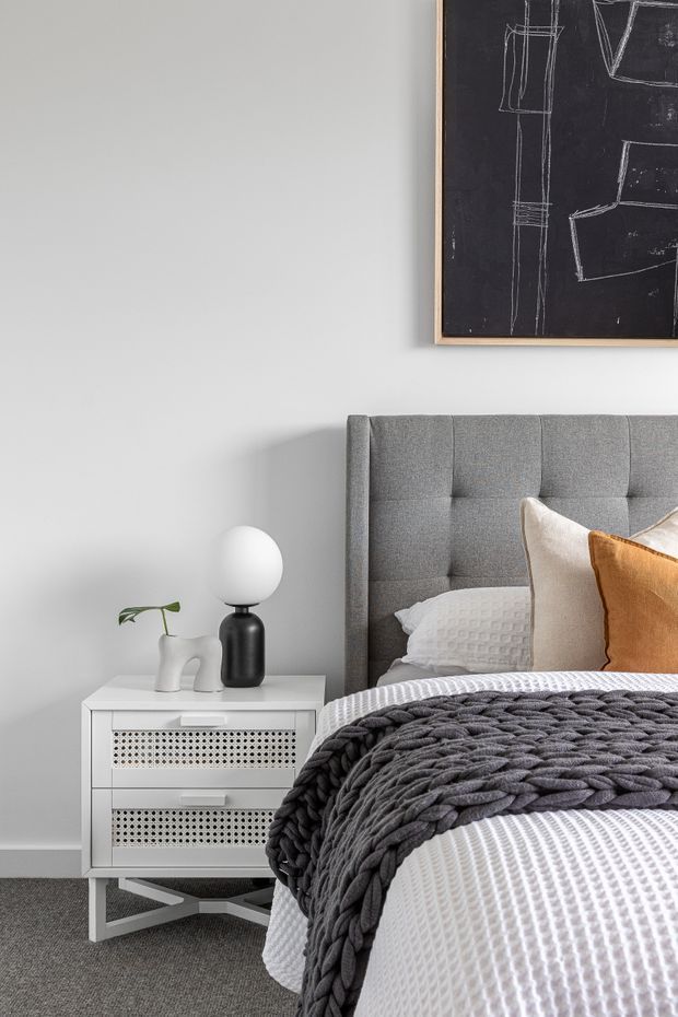 A softly styled contemporary bedroom featuring a grey upholstered, channel-tufted bedhead, white textured bedding, a chunky charcoal knitted throw, and warm ochre and cream cushions. A white perforated bedside cabinet holds a sculptural black-and-white lamp and small plant. The room has pale grey walls, grey carpet, and framed monochrome artwork.