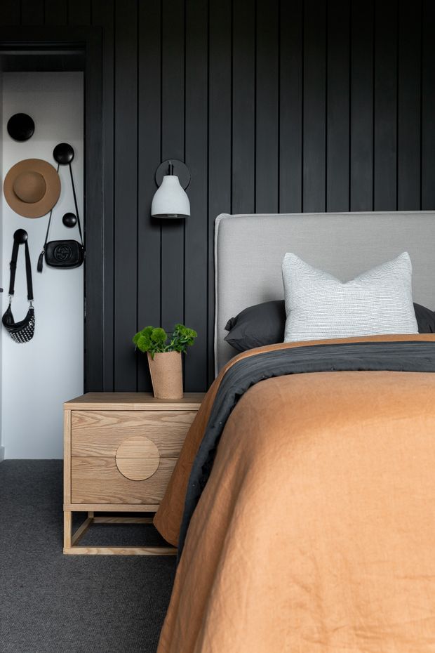 A contemporary bedroom featuring a dark vertical-panel feature wall, a light upholstered bedhead, layered charcoal and tan bedding, and a timber bedside table with rounded drawer details. A wall-mounted pendant and small potted plant add functional and natural accents. An open wardrobe or entry area is visible at left with hanging hats and bags.