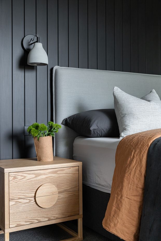 A close-up bedroom interior featuring a large upholstered light-grey headboard and bed, dark vertical timber wall panelling, a wall-mounted grey bedside lamp, and a light timber bedside table with a potted plant. Black and white bedding with a warm brown throw creates a refined, moody atmosphere.