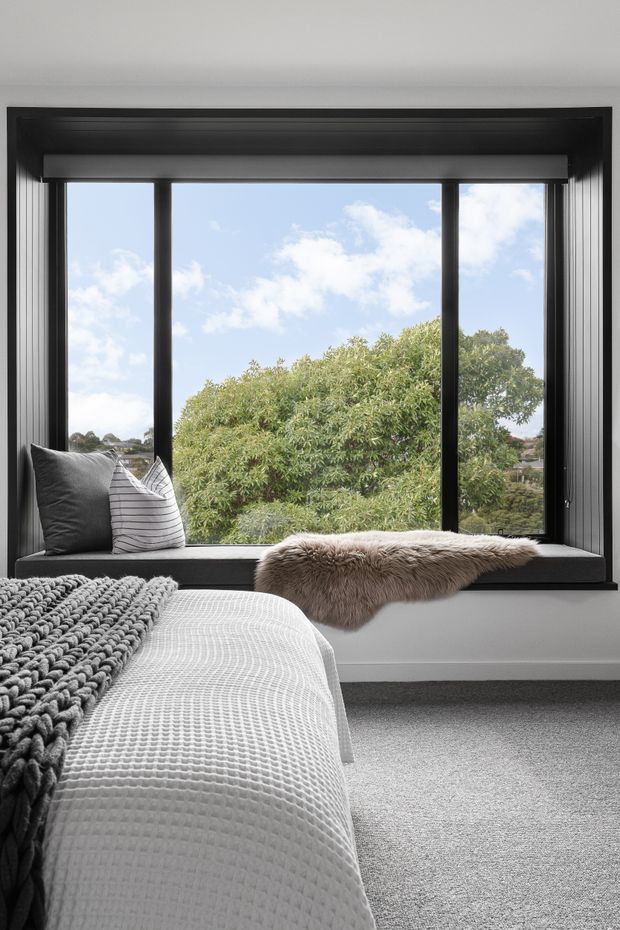 A calm, contemporary bedroom featuring a large black-framed window with a deep built-in sill seat, layered grey and white bedding, textured knitted blanket, striped cushions, and a soft fur throw. The window overlooks leafy greenery and blue sky.