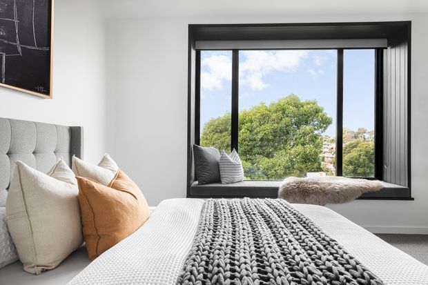 A bright, contemporary bedroom with a large black-framed window and integrated window seat overlooking leafy suburban surroundings. The room features a light upholstered bedhead, neutral bedding, layered cream and tan cushions, and a chunky grey knitted throw. A chalkboard-style artwork is partially visible on the wall.
