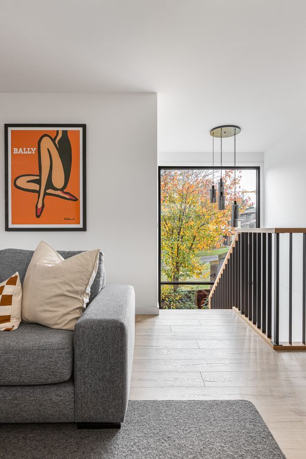 Bright contemporary living area opening onto a stair landing. A large grey sectional sofa sits on a grey rug beside a white wall with framed artwork. Pale timber flooring, a black-and-timber stair balustrade, and a sculptural multi-pendant light define the space, with full-height glazing overlooking autumn foliage.