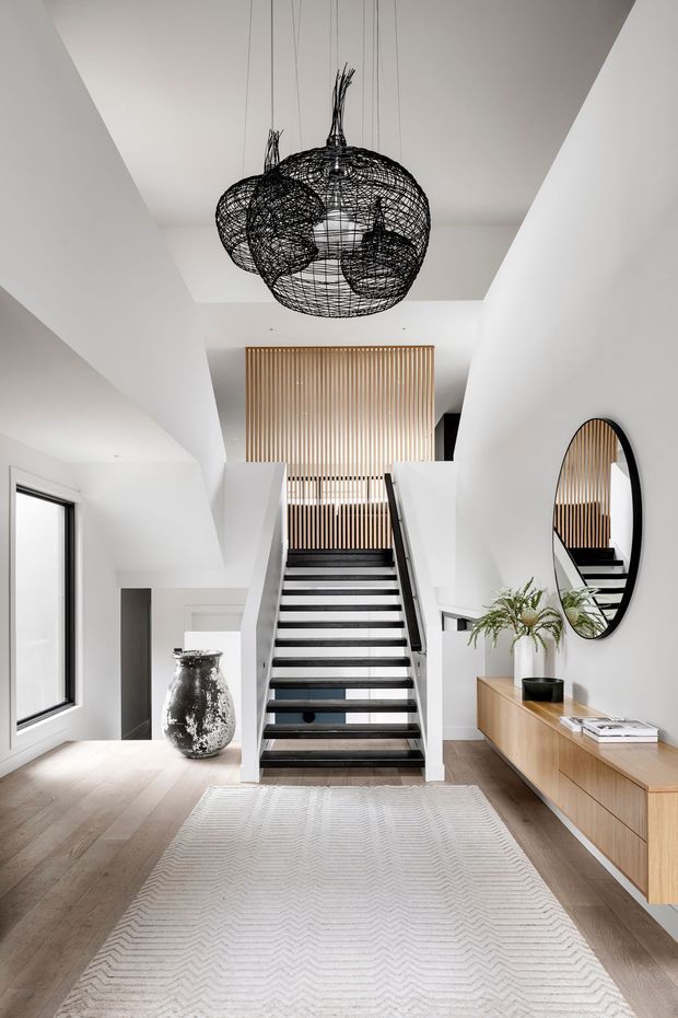 A bright contemporary residential stair hall with a dramatic black wire pendant light, white walls, pale timber flooring, and a central staircase with dark treads and a glass balustrade. Vertical timber slats screen the upper landing, while a timber console, oval black-framed mirror, ceramic vase, plant, and pale patterned rug furnish the entry area.