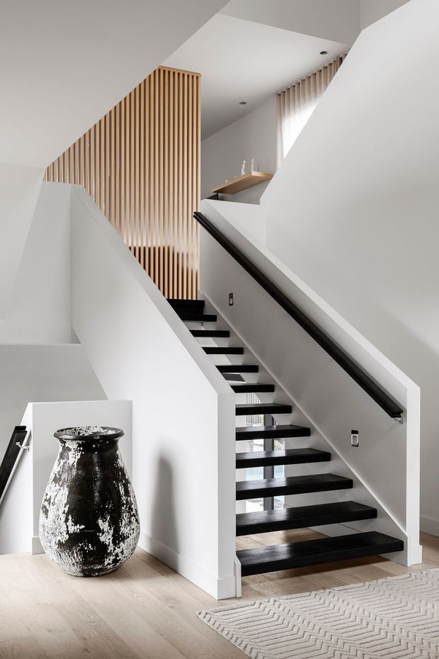 Bright contemporary stairway with floating dark timber treads, white walls and balustrades, and a black handrail. Vertical timber screening rises beside the upper stairwell, with soft daylight filtering through sheer curtains. Pale timber flooring and a textured neutral rug frame a large decorative ceramic vessel in the foreground.