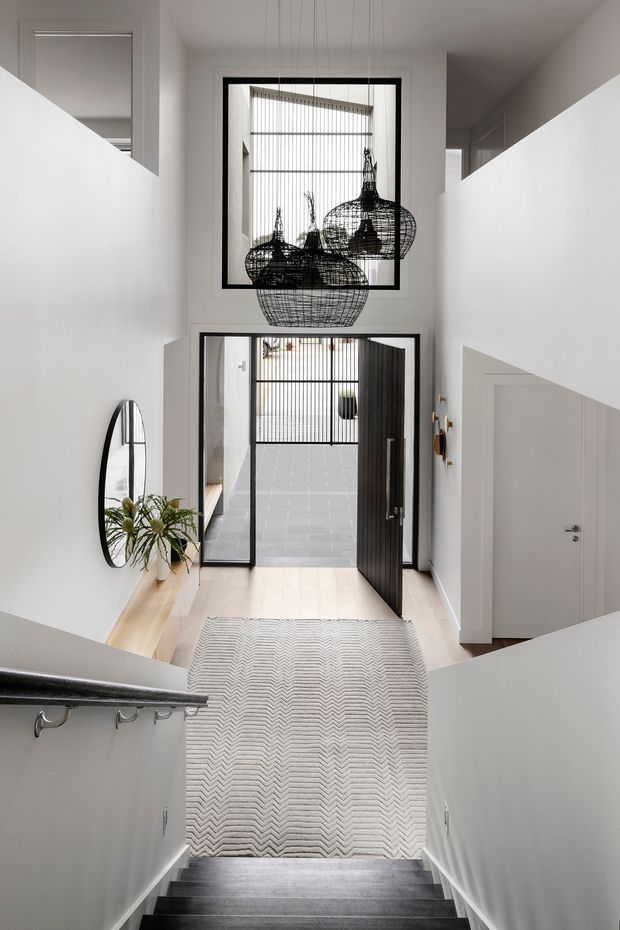 Bright, double-height residential entry hall viewed from a staircase. White walls and angular stair geometry frame a black-framed glazed front door and upper window, with sculptural black wire pendant lights suspended in the void. A pale timber floor, grey stair treads, patterned runner, round wall mirror, and minimal console styling create a crisp contemporary entrance.