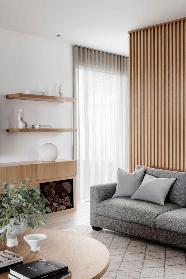 Bright contemporary living room with a grey textured sectional sofa, pale timber oval coffee table, white walls, open timber shelving, built-in firewood storage, sheer curtains, and a prominent vertical timber slat feature wall. Neutral furnishings and abundant filtered daylight create a calm, refined atmosphere.