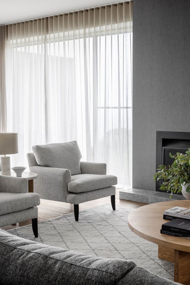 A calm, refined living room with floor-to-ceiling sheer curtains, a large glazed window, grey upholstered armchairs and sofa, a pale patterned rug, and a round timber coffee table. A dark textured feature wall incorporates a contemporary fireplace with a stone hearth, while soft daylight creates an airy neutral atmosphere.