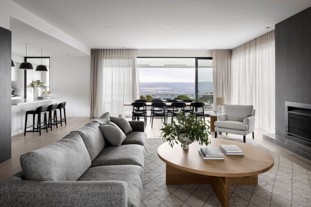 A refined open-plan living and dining area with a contemporary kitchen at left. The room features a large grey sectional sofa, round timber coffee table, light upholstered armchair, dining table and chairs, floor-to-ceiling sheer curtains, and expansive glazing overlooking a distant landscape. A dark fireplace wall anchors the living zone, while recessed lighting and pale finishes create a calm, spacious interior.