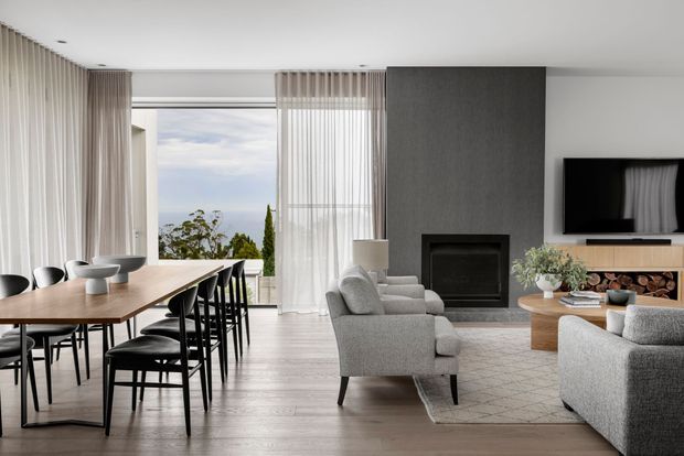 A refined open-plan living and dining room with a neutral contemporary palette. A long timber dining table with black chairs occupies the left side, while grey upholstered armchairs and sofas face a television and built-in fireplace on a dark feature wall. Full-height sheer curtains frame expansive glazing with an outdoor view, and pale timber flooring runs throughout.