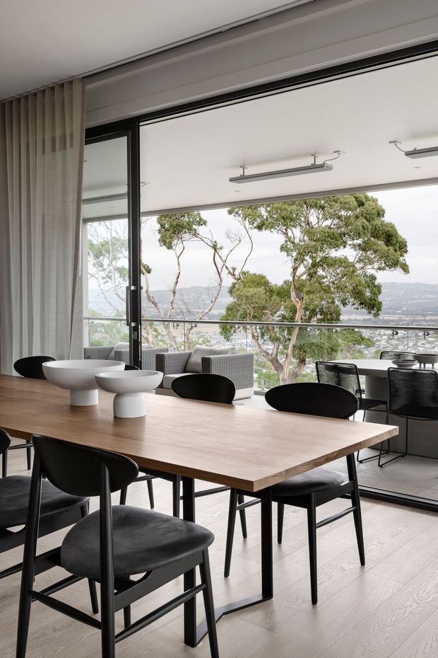 A bright, contemporary dining area with a large light timber dining table, black upholstered dining chairs, and sculptural white tabletop bowls. Full-height glazed doors open to a balcony or terrace, with a green, hilly landscape visible beyond. The space features pale timber flooring, neutral curtains, black window frames, and linear ceiling-mounted lighting.