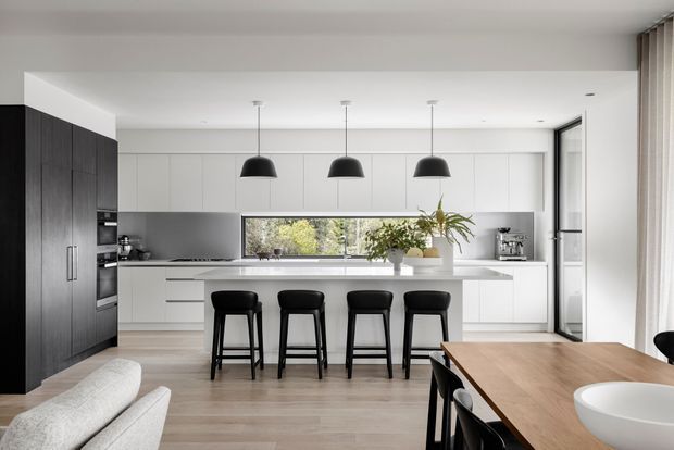 Bright contemporary open-plan kitchen and dining area with a long white island, four black pendant lights and four black bar stools. The kitchen features extensive white flat-panel cabinetry, a dark timber-toned tall cabinet wall, integrated appliances, pale timber flooring, and a horizontal window overlooking greenery. A timber dining table and upholstered seating occupy the foreground.