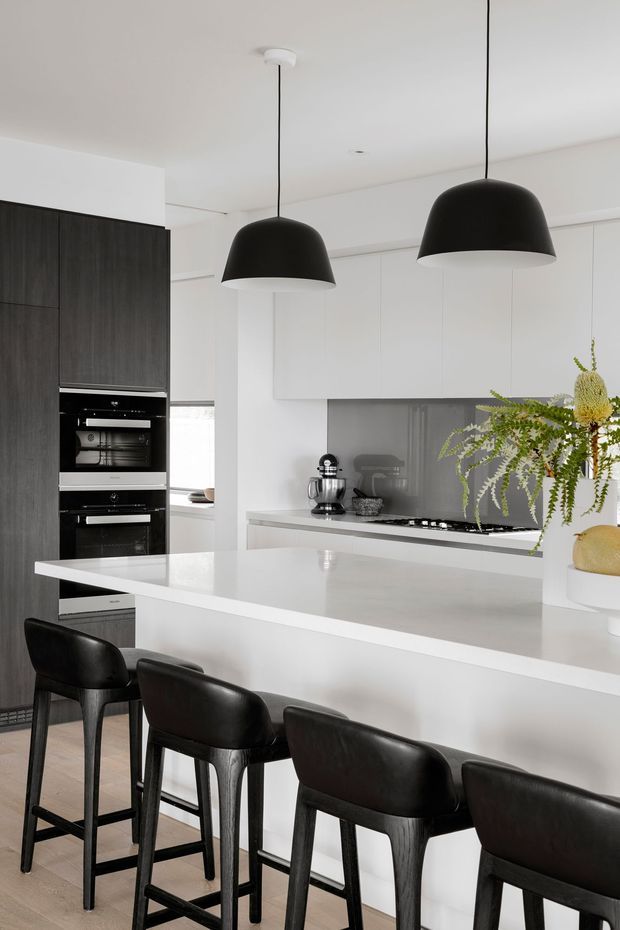 A bright, contemporary kitchen with a large white island and integrated breakfast bar seating. The space features dark timber-look full-height cabinetry with built-in ovens, white flat-panel overhead cabinetry, a grey reflective splashback, a pale benchtop, black dome pendant lights, and several dark upholstered bar stools. A potted fern adds greenery to the otherwise monochromatic scheme.