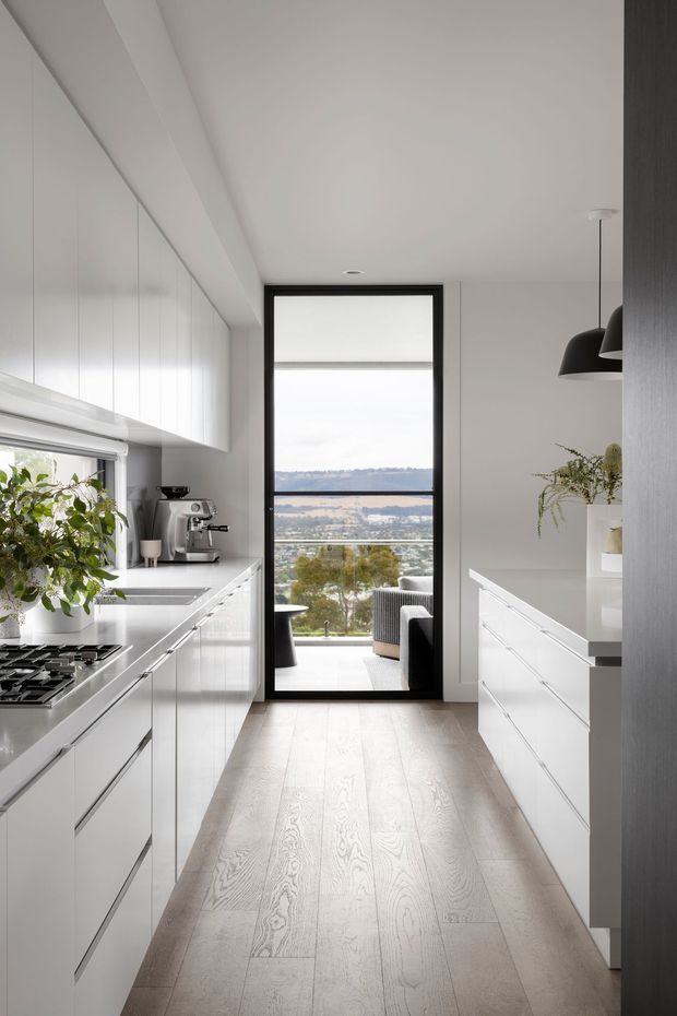 A bright, narrow contemporary kitchen with white floor-to-ceiling cabinetry on both sides, integrated appliances, a gas cooktop, sink, coffee machine and pale timber flooring. Black-framed full-height glazing at the end opens to a balcony or terrace with a distant landscape view. Minimal black pendant lights and dark accents contrast with the predominantly white interior.